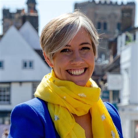 Daisy Cooper of the Liberal Democrates.  A Smiling adult with short blonde hair wearing a bright yellow scarf and blue jacket, standing outdoors in front of white timbered houses and a stone building.